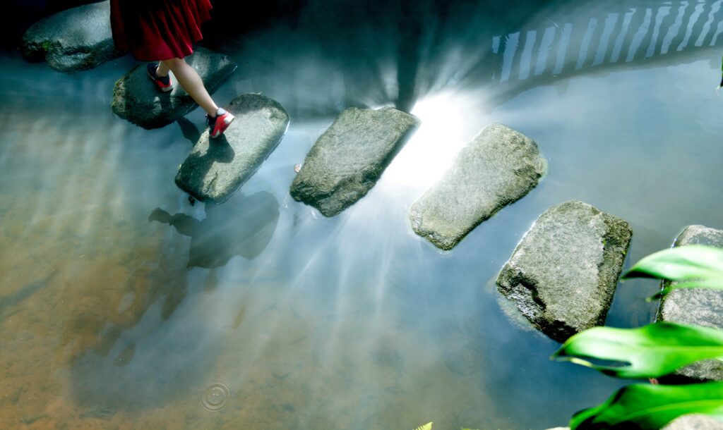 A woman crosses a river over rocks with reflection of a bridge in the water.